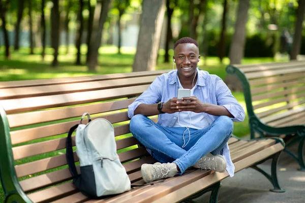 Прохолодний чорний хлопець в навушниках прослуховування музики онлайн зі свого смартфона на лавці в парку, порожній простір — стокове фото
