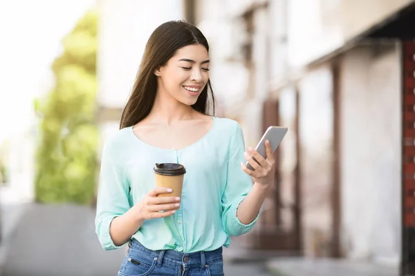快乐的亚洲女孩，在户外用智能手机喝着外卖咖啡，浏览社交媒体 — 图库照片