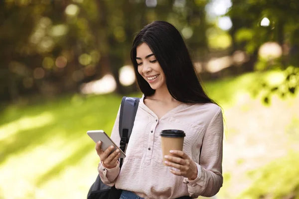 Sırt çantası ve kahveyle gezerken akıllı telefon kullanan Asyalı öğrenci. — Stok fotoğraf