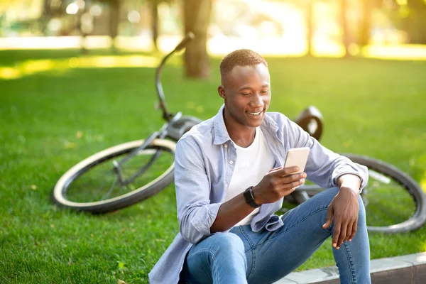 Веселий афроамериканець з велосипедом, який переглядає Інтернет на своєму смартфоні в міському парку. — стокове фото