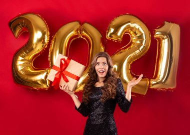 Yeni yıl 2021. Zarif elbiseli, kırmızı arka planda altın balonlu hediye kutusu taşıyan genç bir kadın.