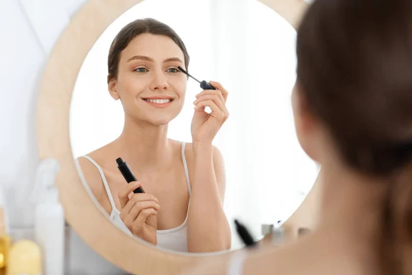 Sorrindo jovem mulher olhando para o espelho, usando rímel — Fotografia de Stock