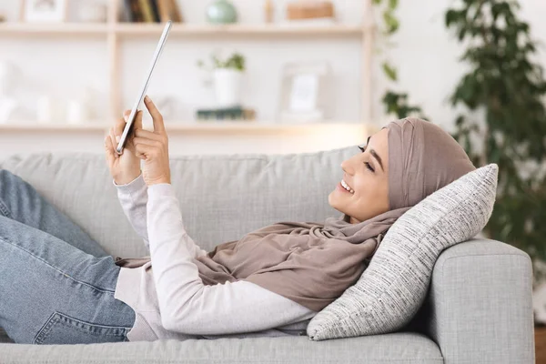 Pasatiempo en casa. Árabe chica en hijab relajarse en el sofá con la tableta digital — Foto de Stock