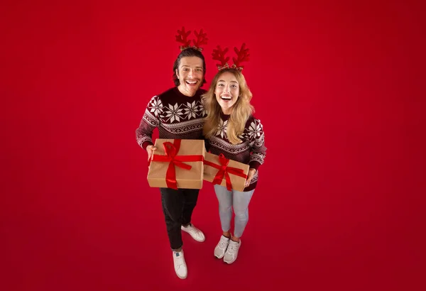 Pareja feliz usando suéteres de lana y cuernos de ciervo con regalo de Navidad sobre fondo rojo — Foto de Stock