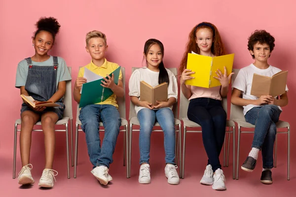 Multinacionális gyerekek tankönyvekkel és másolatokkal székeken ülve, rózsaszín háttér felett kamerával — Stock Fotó