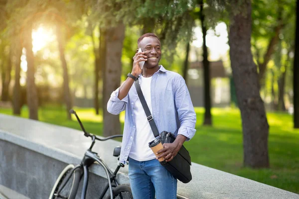 Młody czarny mężczyzna z kawą na wynos stojący obok roweru w parku i rozmawiający przez komórkę — Zdjęcie stockowe
