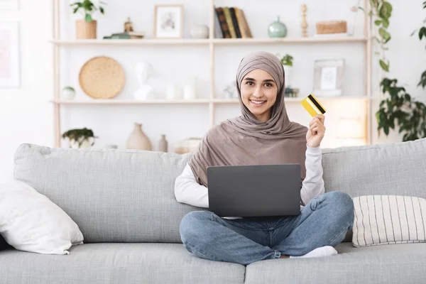 Transferencias de dinero en línea. Sonriente chica árabe usando el ordenador portátil y la tarjeta en casa — Foto de Stock
