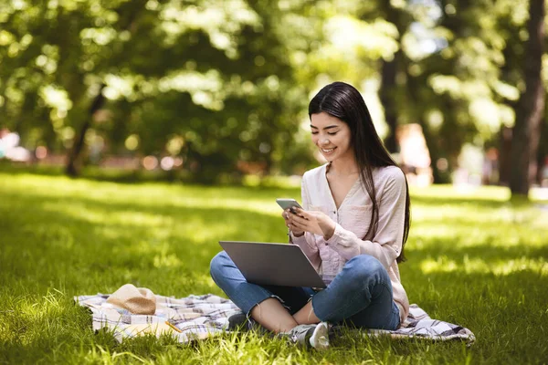 Fröhliche Koreanerin entspannt sich mit Smartphone und Laptop im Freien und sitzt im Park — Stockfoto