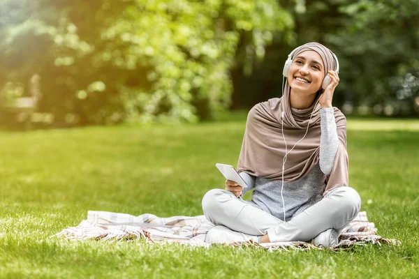 Mujer feliz en hijab disfrutando de la música mientras descansa en el parque — Foto de Stock