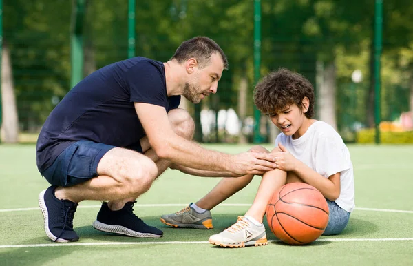 Teacher helping boy with knee trauma after playing basketball — Stock Photo, Image