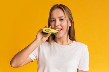 Avokado 'nun yarısını ısıran ve kameraya gülümseyen mutlu genç kadın.