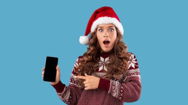 İnternetten Noel alışverişi. Şok olmuş genç kız, Noel Baba şapkasıyla cep telefonunu işaret ediyor, boş ekran, mavi arka plan