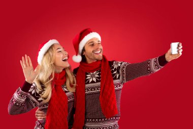 Noel eğlencesi. Noel Baba şapkalı romantik genç çift kırmızı arka planda selfie çekiyorlar.