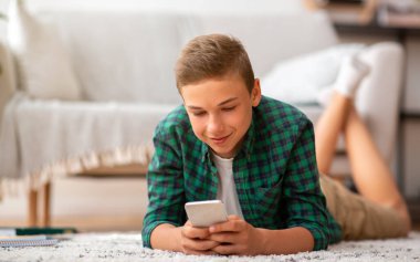 Neşeli genç adam evde yerde uzanıyor, telefon kullanıyor.