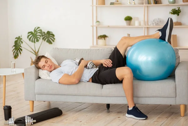 Lenistwo i trening. Zmęczony facet śpiący na kanapie z hantlami i piłką fitness — Zdjęcie stockowe