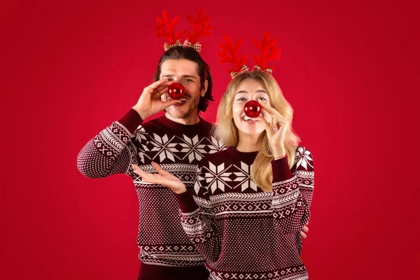 Diversión de Navidad. Tonta joven pareja sosteniendo bolas de Navidad cerca de sus narices sobre fondo rojo — Foto de Stock
