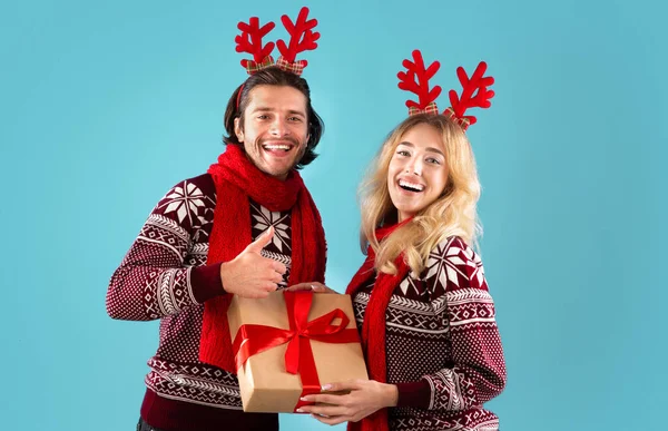 Romántica pareja joven en trajes de Navidad sosteniendo caja de regalo y mostrando el gesto de pulgar hacia arriba sobre fondo azul — Foto de Stock