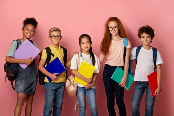 Bereit für die Schule. Kinder verschiedener Nationalitäten mit Notizbüchern und Rucksäcken blicken vor rosa Hintergrund in die Kamera — Stockfoto