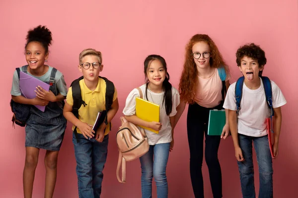 Глупые мультиэтнические дети со школьными принадлежностями делают смешные лица на розовом фоне — стоковое фото