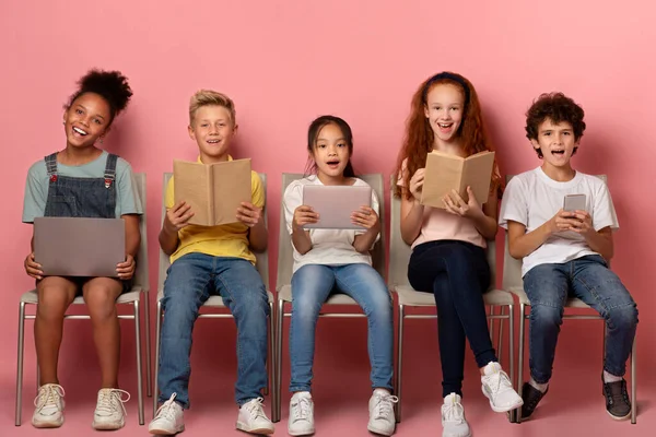 Enfants excités avec des livres et des gadgets pour l'éducation à distance assis sur des chaises sur fond de studio rose — Photo