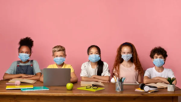病毒爆发期间的教育。不同的学童戴着医疗面具坐在桌子前，粉红的背景，有空余的空间 — 图库照片