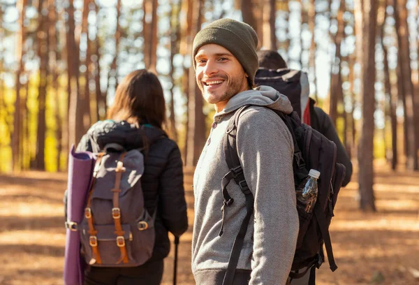 Usmívající se mladý muž batohem turistika s přáteli v lese — Stock fotografie