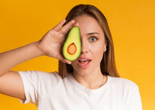 Ung flicka som håller Avokado halvnära ansikte, täcker ögat — Stockfoto