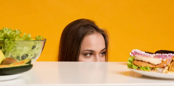 Donna che sbircia fuori dalla tavola, scegliendo tra cibo sano e malsano — Foto Stock