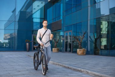 Çantalı ve kahveli adam, bisikletle ofis binasının yakınına gidiyor.