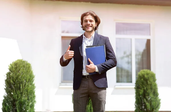 Positivo joven agente inmobiliario con documentos que muestran el gesto de pulgar hacia arriba cerca de la casa para la venta al aire libre — Foto de Stock