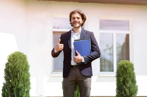 Positiver junger Makler mit Dokumenten, die Daumen-hoch-Geste in der Nähe von Haus zum Verkauf draußen zeigen — Stockfoto