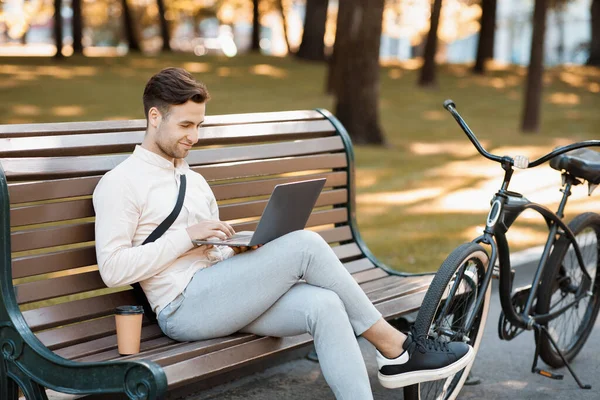 Trabajo durante el descanso al aire libre. Tipo atractivo se sienta en el banco, tipos en el ordenador portátil con café y bicicleta — Foto de Stock