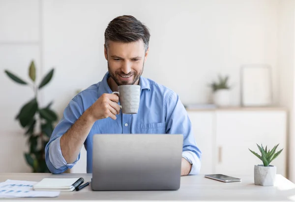 Dia casual no escritório. Sorrindo masculino empregado usando laptop e beber café — Fotografia de Stock