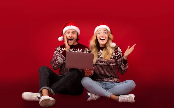 Heyecanlı genç çift, kırmızı arka planda dizüstü bilgisayarda süper indirimli Noel hediyeleri alıyor. — Stok fotoğraf