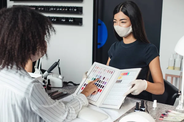 Menina asiática em máscara protetora olha para o cliente americano africano, escolhe a cor no interior do estúdio de unhas — Fotografia de Stock