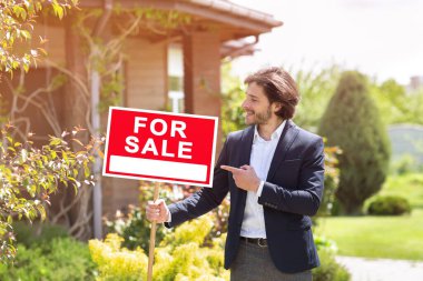 Güvenilir bir emlakçı, dışarıdaki apartmanın önünde satılık sözlerle tabelayı işaret ediyor.