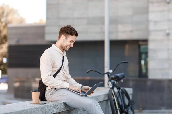 Freelancer werkt buiten. Glimlachende man in hoofdtelefoon met koffie surfen in laptop — Stockfoto