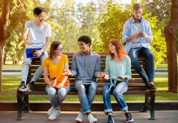Estudiantes multiraciales se relajan después de clases sentados en el banco en el parque — Foto de Stock