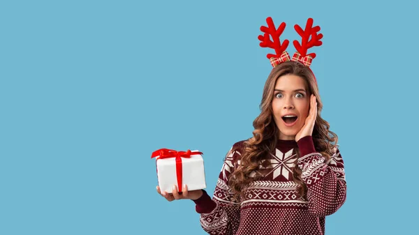 Shopping per Natale. Sorpresa giovane signora in maglia maglione che mostra scatola regalo su sfondo blu, spazio per il testo — Foto Stock