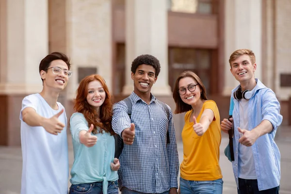 Různí studenti gestikulující palce nahoru s úsměvem na fotoaparát stojící venku — Stock fotografie