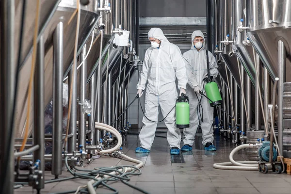 Nettoyage et désinfection. Deux hommes en combinaison de protection et sac de pulvérisation travaillent à l'usine — Photo