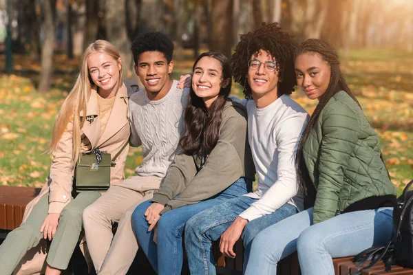Grupo de jóvenes internacionales sentados en el banco del parque público —  Fotos de Stock