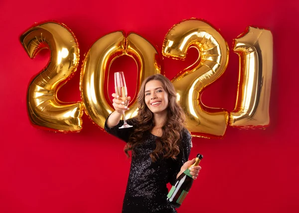 Célébrons 2021. Belle jeune femme avec bouteille et verre de champagne sur fond rouge avec des ballons en or — Photo