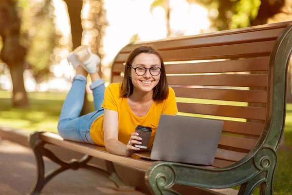 Chica estudiante feliz usando el ordenador portátil acostado en el banco al aire libre — Foto de Stock