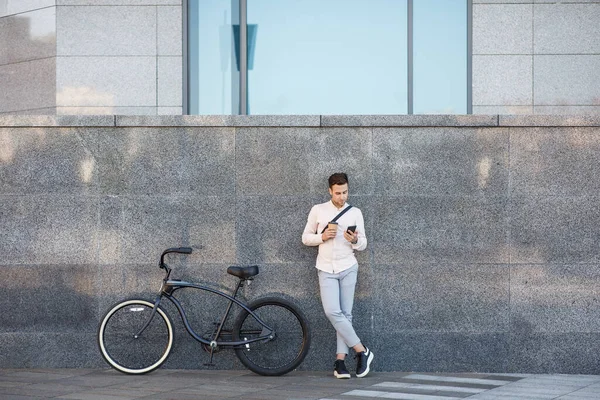 Online ao ar livre. Empresário com bicicleta e xícara de café, olha para o smartphone encostado à parede — Fotografia de Stock