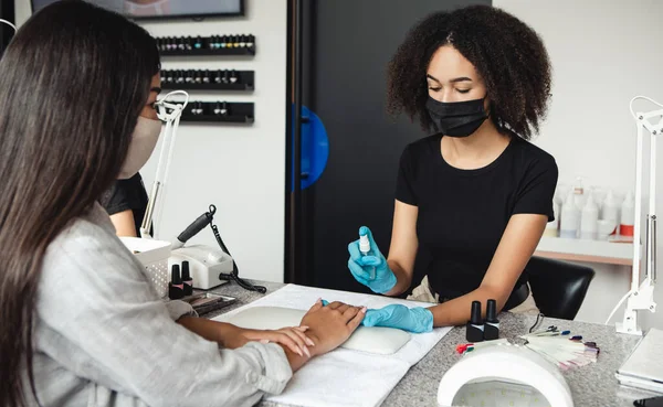 Nagelsalon tijdens quarantaine. Afrikaans amerikaans meisje in beschermende masker desinfecteert handen van cliënt — Stockfoto