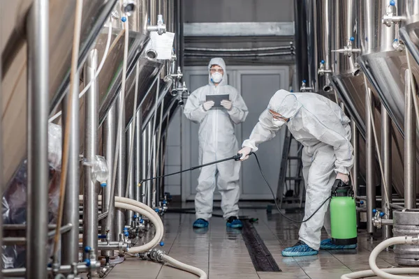 Calidad del producto y desinfección. Los hombres en trajes de materiales peligrosos trabajan con bolsas de spray en la fábrica de cerveza — Foto de Stock