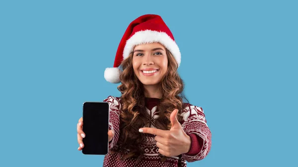 Attractive young girl in Christmas outfit pointing at smartphone with blank screen on blue background, mockup for design — Stock Photo, Image