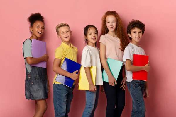 Niños y niñas alegres de diferentes nacionalidades con copybooks posando sobre fondo rosa — Foto de Stock