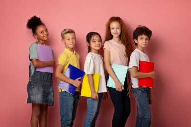 Ellerinde not defterleriyle pembe arka planda duran neşeli çoklu etnik kökenli öğrenciler.